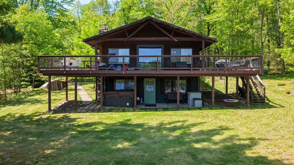 Rustic log cabin with large wooden deck surrounded by lush green trees and manicured lawn.