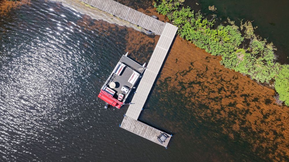 Aerial view of a boat docked at a wooden pier on a calm lake surrounded by lush green and brown vegetation.