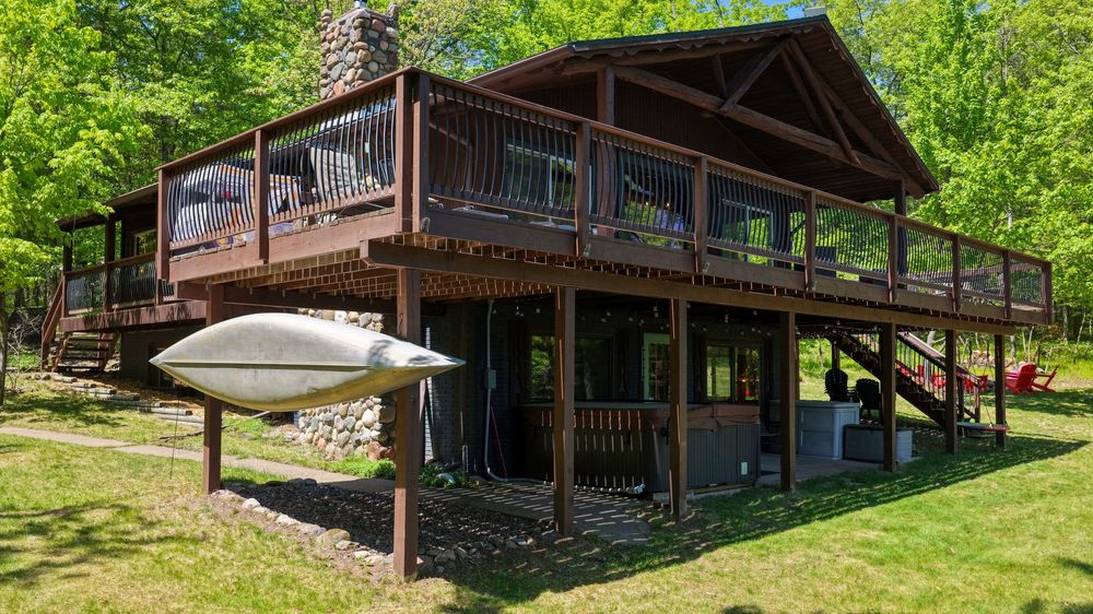Rustic wooden cabin with large deck, stone chimney, kayak storage, and lush green trees in a serene wooded setting.