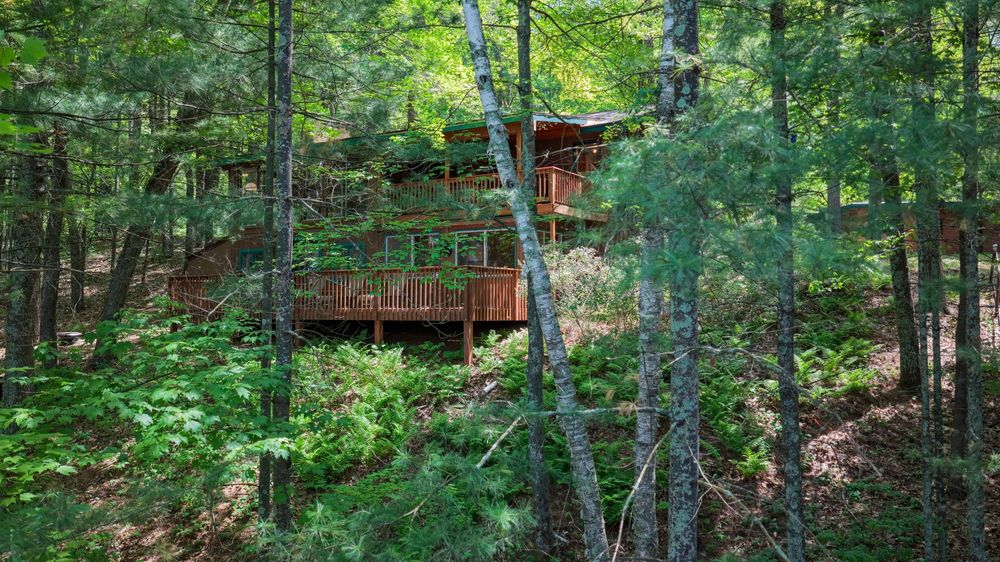 Rustic wooden cabin nestled among lush green forest trees, featuring a large deck surrounded by tall pines.