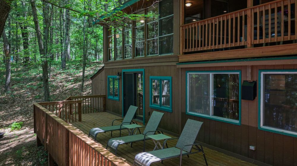 Rustic two-story cabin exterior with teal-trimmed windows, wooden deck, lounge chairs, and lush forest surroundings.