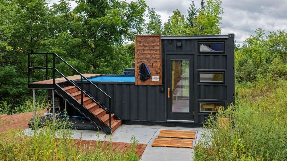 Modern shipping container home with rooftop pool, wooden staircase, and glass doors surrounded by lush green trees.
