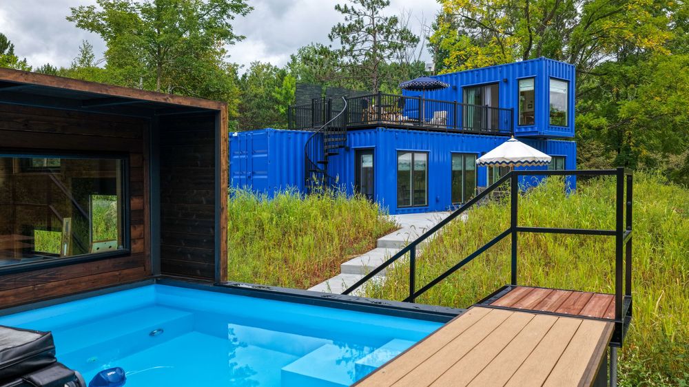 Blue shipping container home with outdoor pool and wooden deck surrounded by lush green trees and meadow.