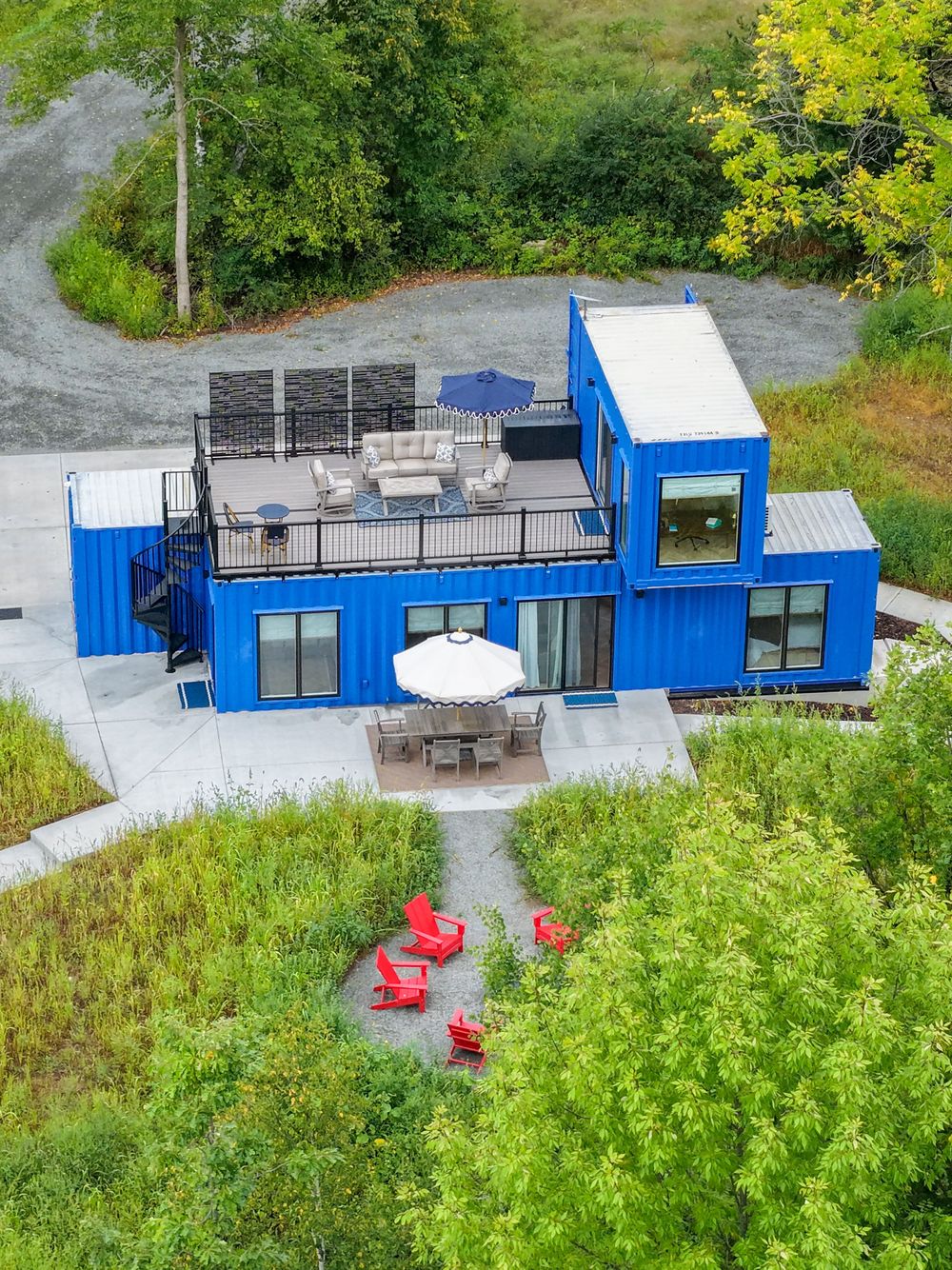 Aerial view of a modern blue shipping container home with rooftop deck, red Adirondack chairs, and outdoor dining area.