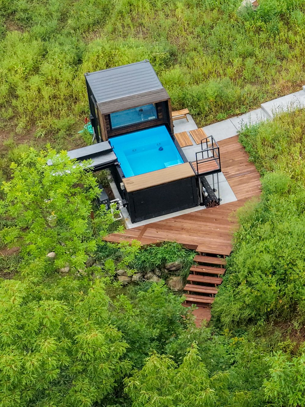Aerial view of a modern container home with a blue swimming pool surrounded by lush green jungle vegetation and wooden deck.