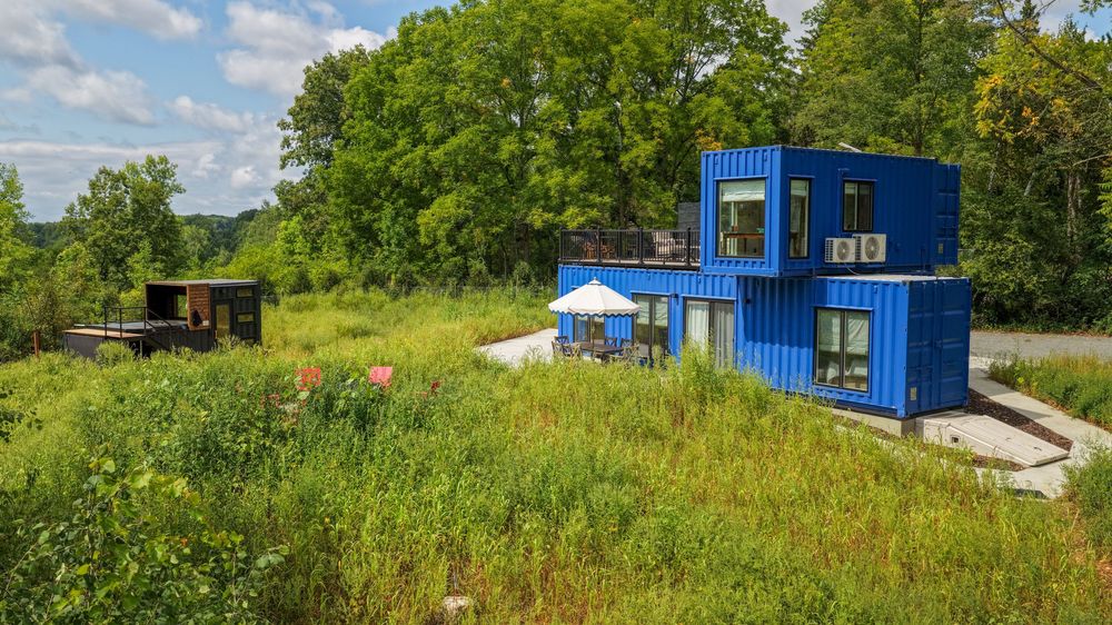 Blue shipping container home surrounded by lush green meadow and trees on a sunny day.