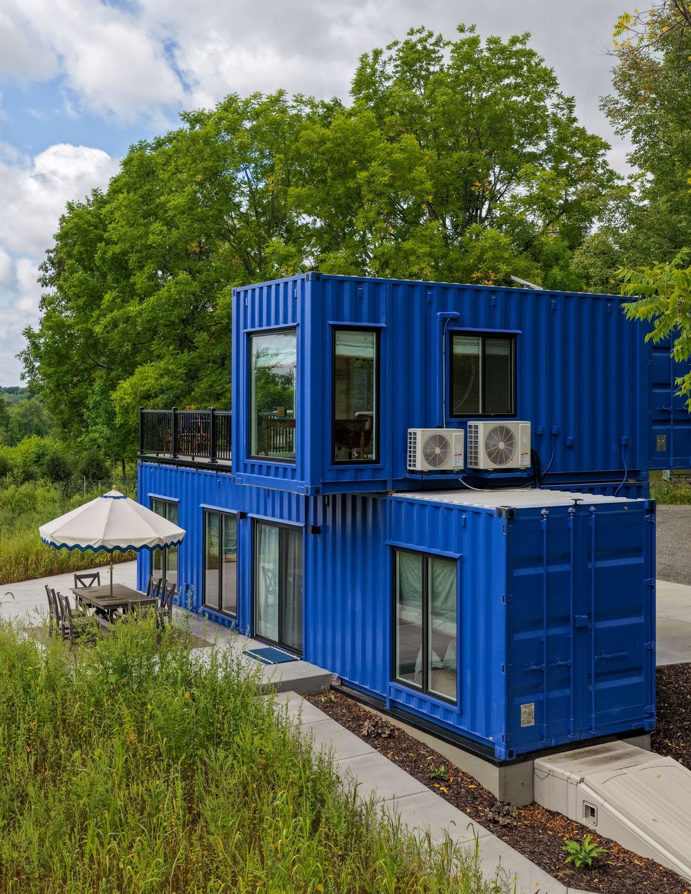 Two-story blue shipping container home with large windows, rooftop deck, and AC units surrounded by lush green trees.