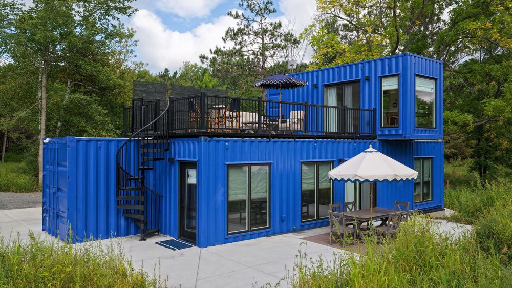 Modern two-story blue shipping container home with large windows, outdoor patio, spiral staircase, and rooftop deck surrounded by trees.