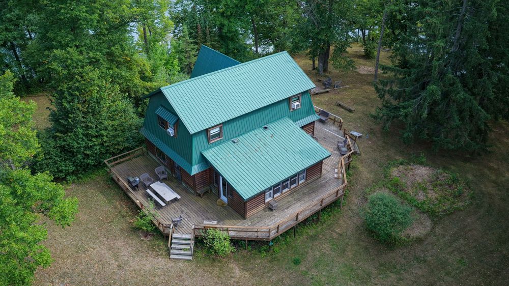 Aerial view of a charming cabin with teal metal roof surrounded by lush green trees and a wraparound wooden deck.