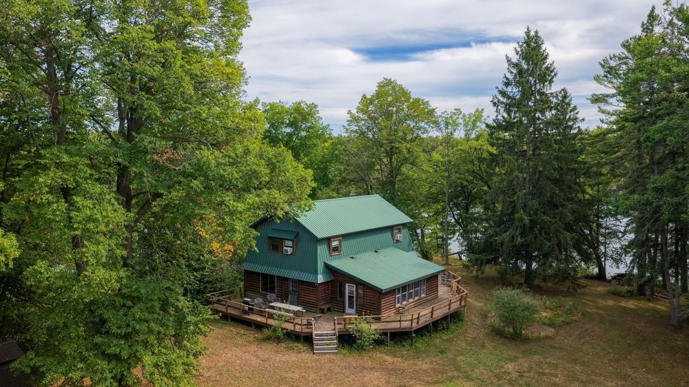 Aerial view of a rustic wooden cabin with green metal roof surrounded by lush trees near a lakeside setting.