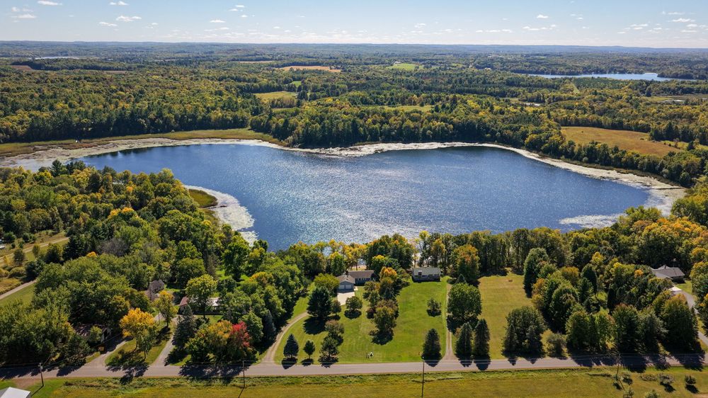 Aerial view of a calm blue lake surrounded by dense autumn forest, with homes and green lawns along the shoreline under a clear sky.