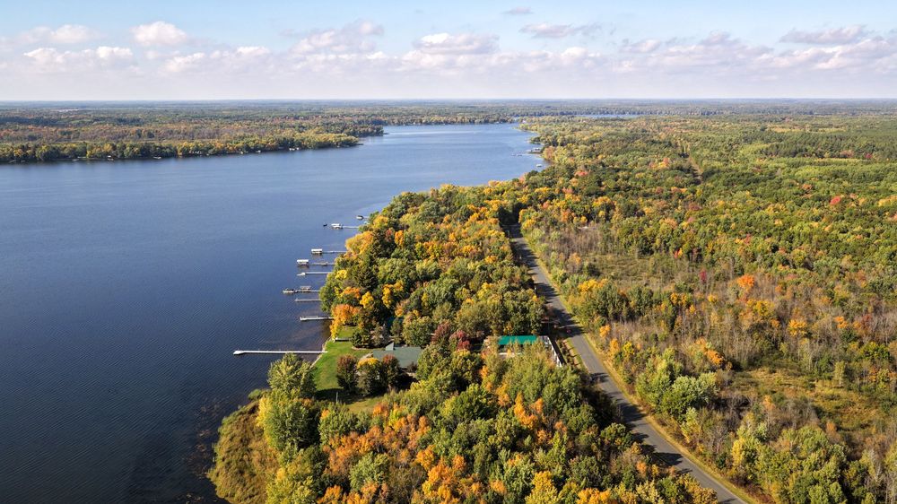 Aerial view of a calm blue lake bordered by colorful autumn forest with a winding road and small docks along the shoreline.
