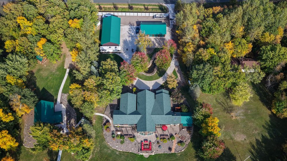 Aerial view of a green-roofed estate with a main house, outbuildings, and a circular driveway surrounded by colorful autumn trees.