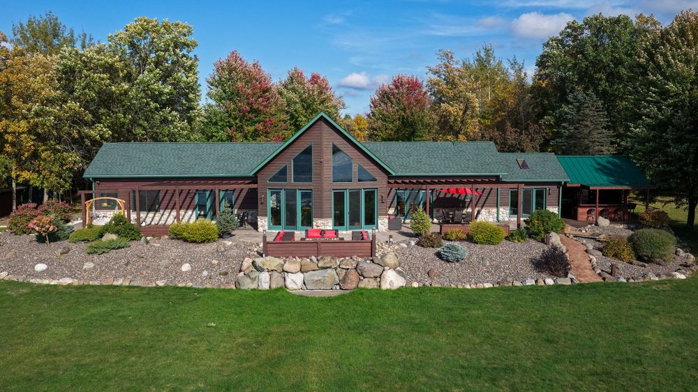 Green-roofed lakefront home with a tall glass gable entry, stone landscaping and a manicured lawn surrounded by colorful autumn trees.