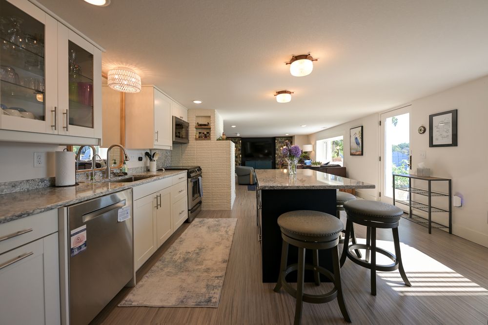 Open-concept kitchen with white cabinets, stainless steel dishwasher, granite counters, and a dark island with two cushioned bar stools.