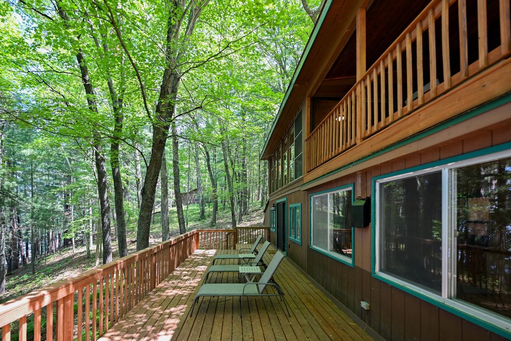 Spacious wooden deck with lounge chairs surrounded by lush green trees beside a rustic cabin with large windows.