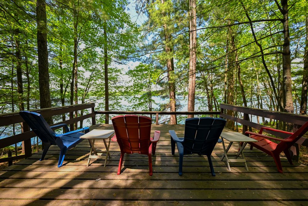 Colorful Adirondack chairs on a wooden deck overlooking a serene lake through tall green forest trees.