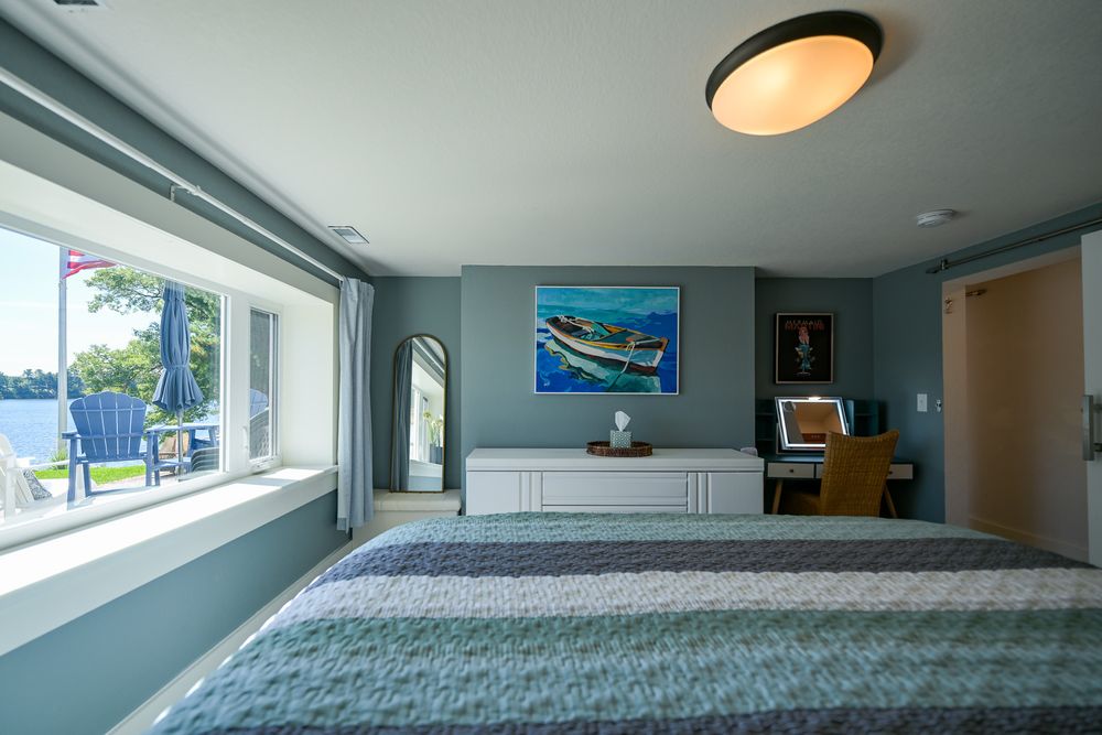 Bright waterfront bedroom with a striped bed, large lakeview window, white dresser, boat painting, and a desk with chair.