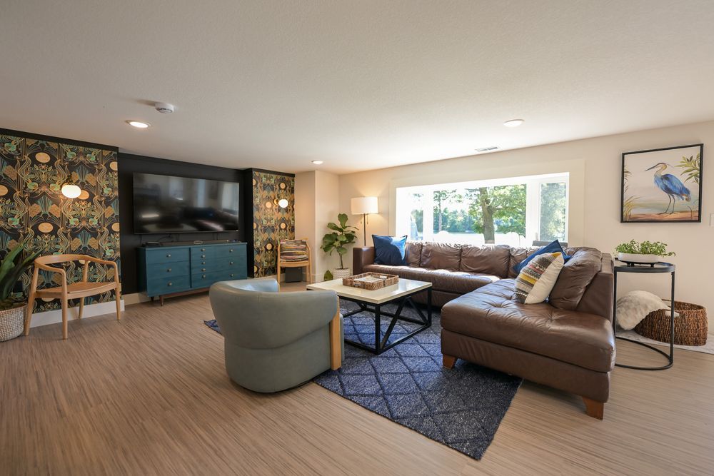 Spacious modern living room with a brown leather sectional, floral accent wall, wall-mounted TV, teal media console and large window.