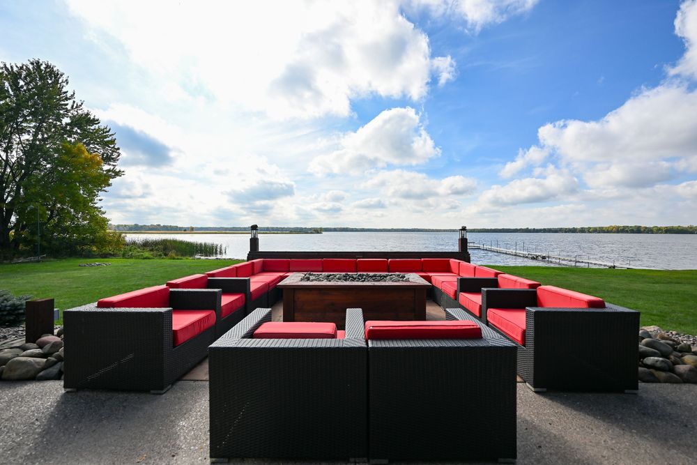 Outdoor lakeside patio with black sectional sofas, red cushions and a central fire pit overlooking calm water under a bright cloudy sky.