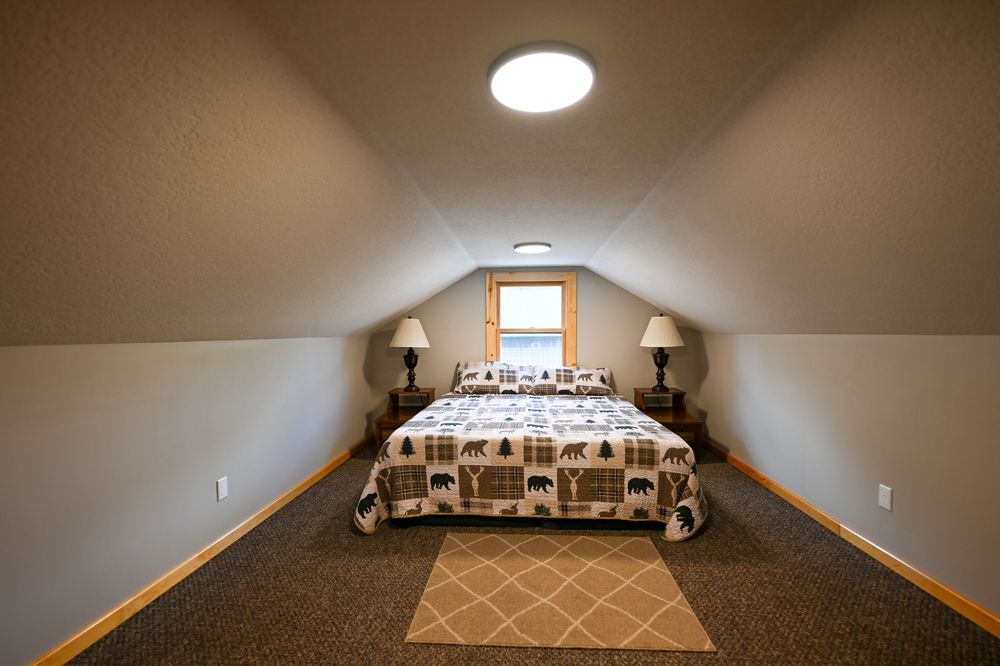 Cozy attic bedroom with sloped ceilings, a patterned quilt on a bed, two bedside lamps, a small window, and a geometric area rug.