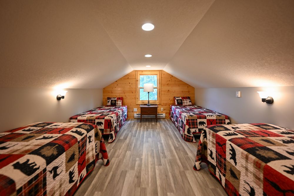 Cozy attic bedroom with four twin beds in rustic bear and moose print quilts, wood plank accent wall, and a window with a small desk.