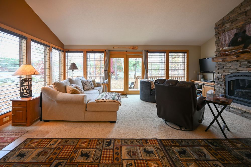 Cozy living room with a beige sectional sofa, stone fireplace, leather recliner, and large windows with a patterned area rug.