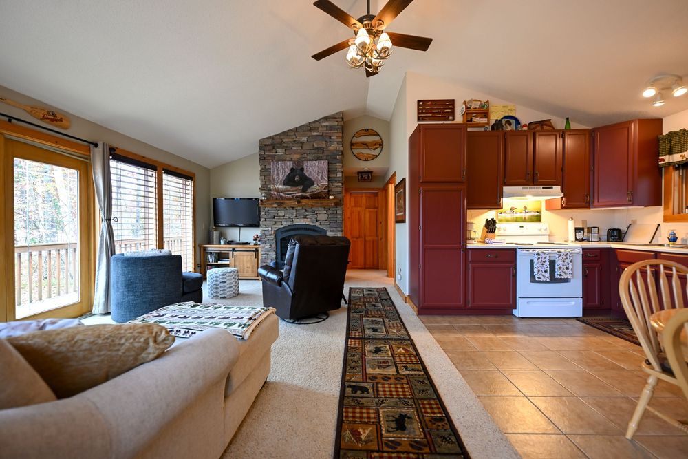 Open-concept cabin living room with vaulted ceiling, stone fireplace, cozy seating, patterned runner rug and a kitchen with red cabinets.