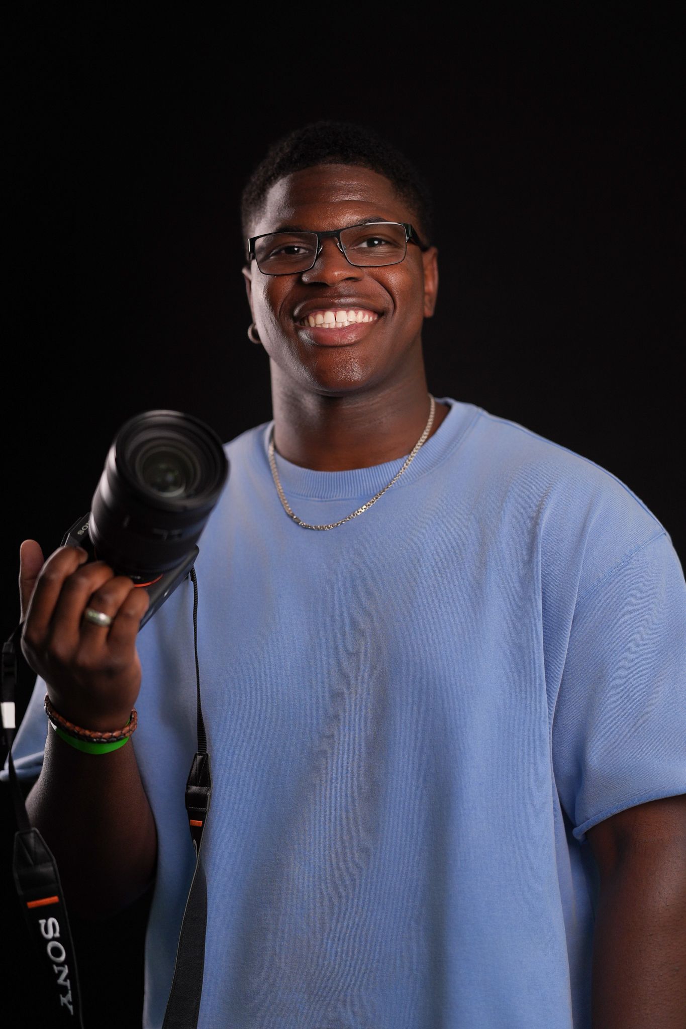 Smiling young man in glasses and a light blue t-shirt holds a Sony camera against a dark background.