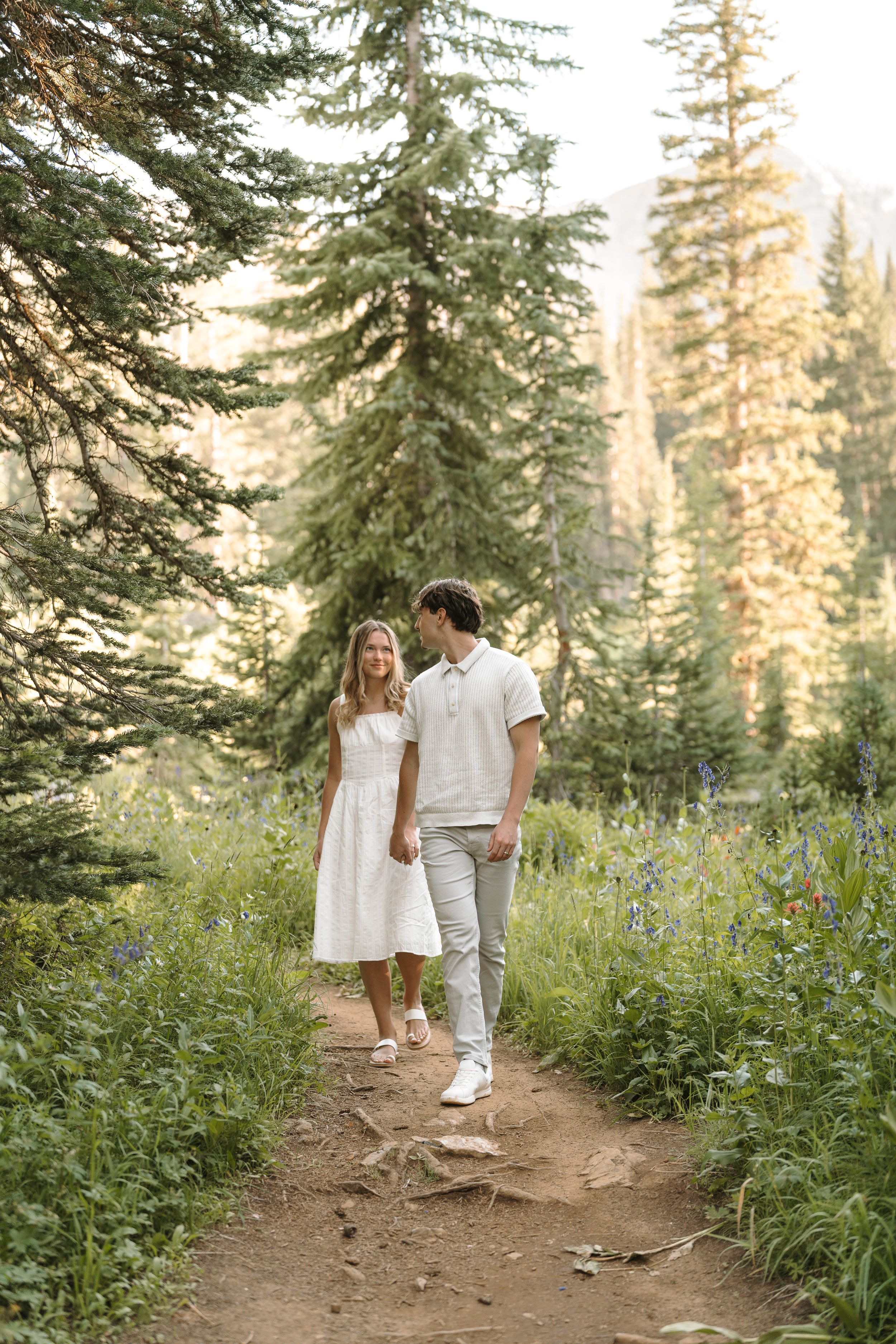 Pair walking hand in hand along forest trail lined with lupines and evergreen trees.