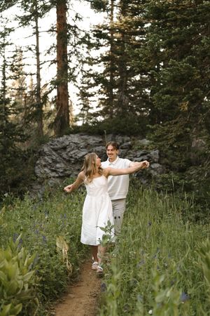 Couple dancing playfully together on a mountain trail surrounded by wildflowers and evergreen trees at sunset.