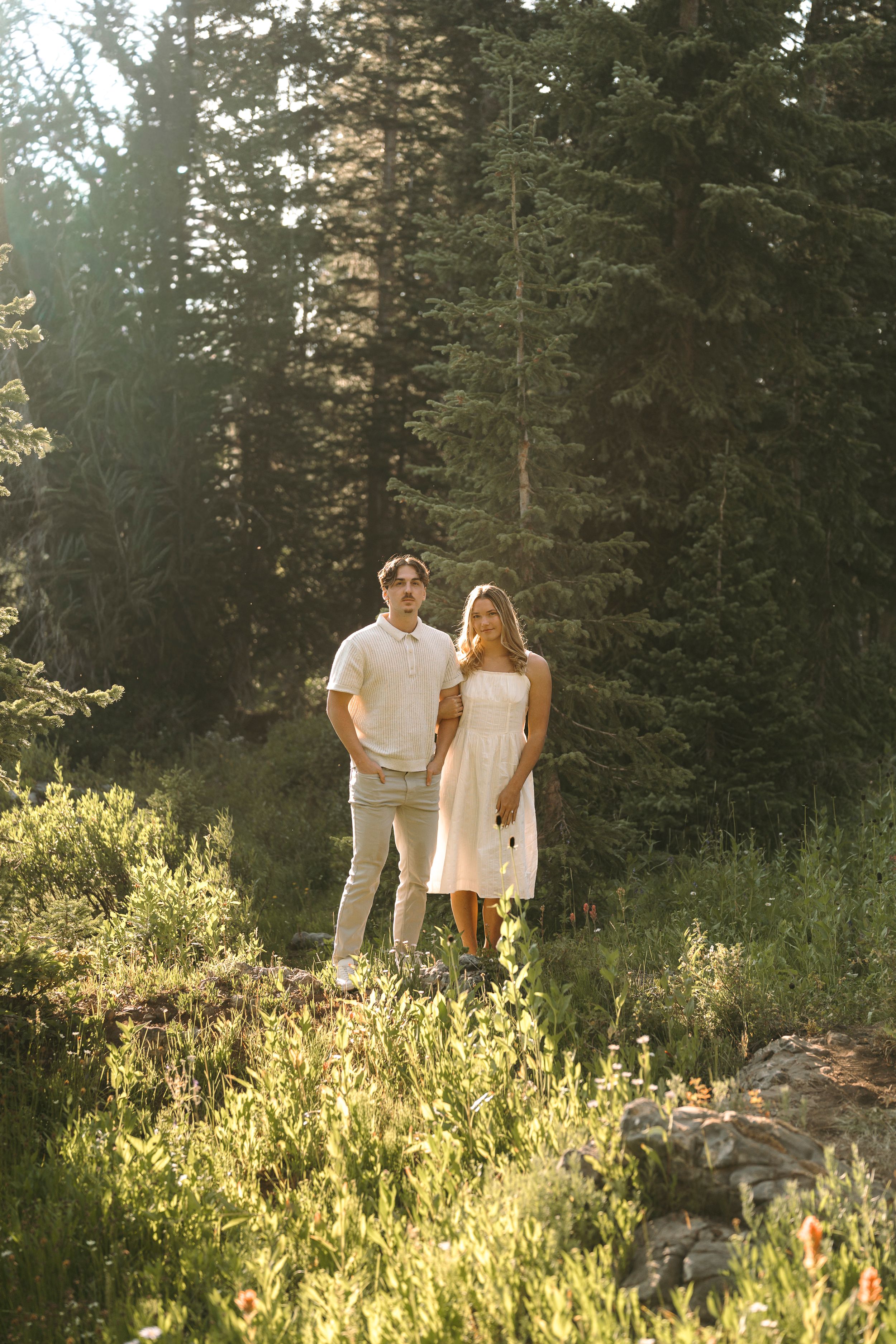 A couple shares an intimate moment in a sunlit mountain clearing surrounded by evergreens.