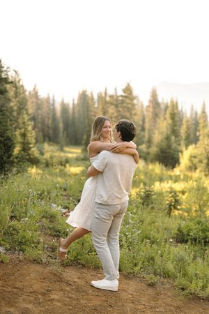 People in white outfits dancing joyfully among wildflowers with sunlit mountain forest in the background.