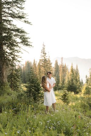Romantic couple in white embraces in a mountain meadow among evergreen trees at sunset.