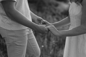 Black and white photo showing close up of two people holding hands with wedding rings outdoors.