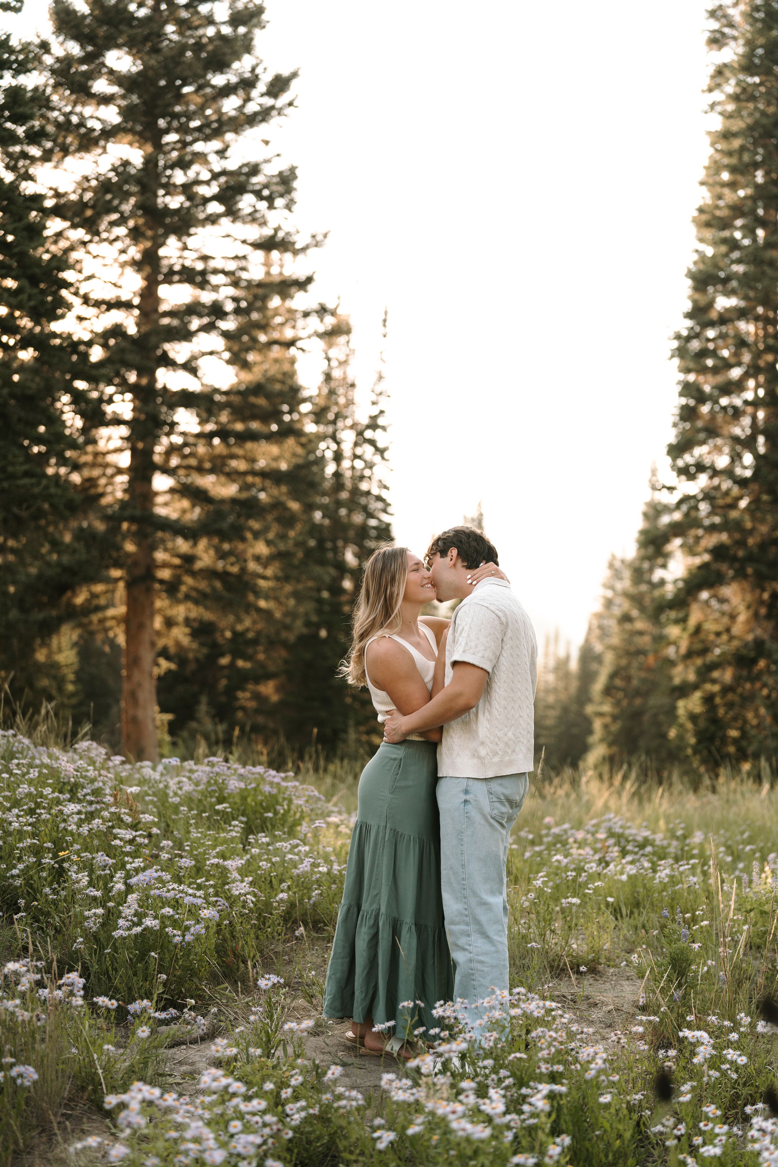 Couple embraces in a wildflower meadow surrounded by pine trees at sunset.