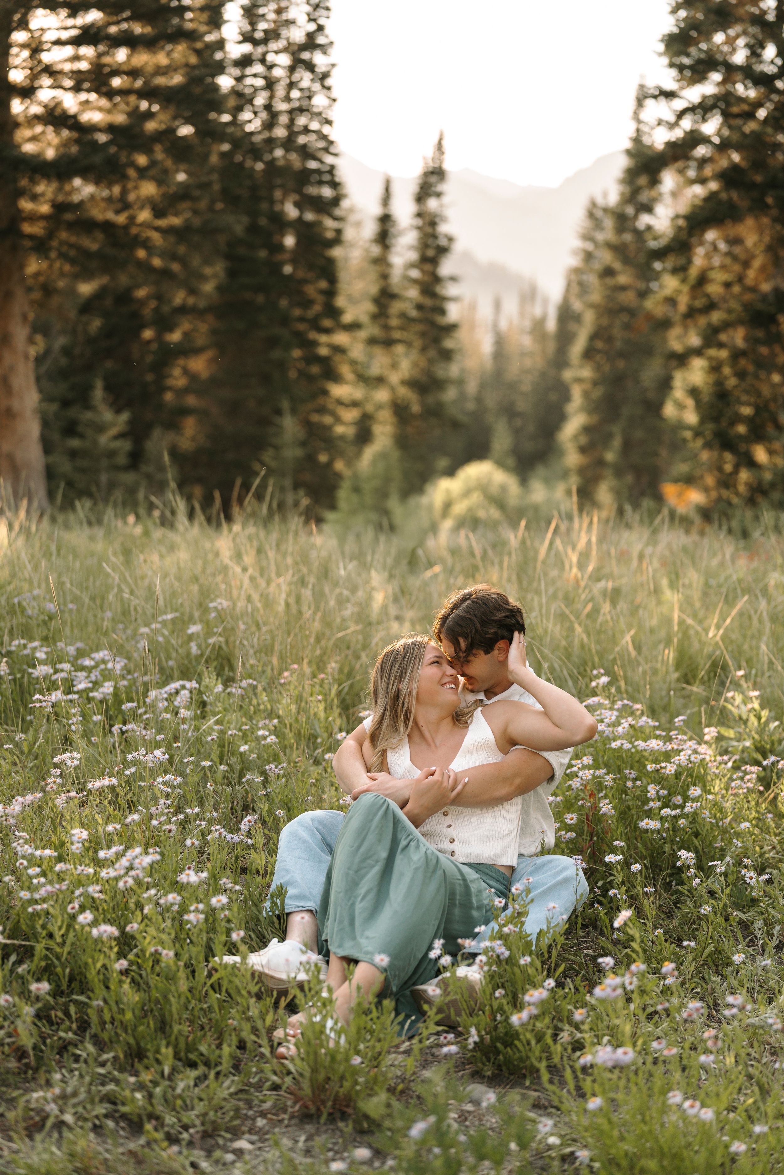 A romantic couple lounges in a wildflower meadow surrounded by evergreen trees at sunset.