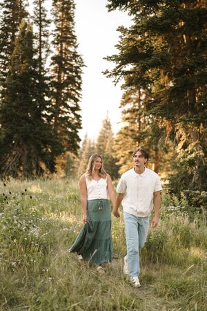 A couple walks hand in hand through a mountain meadow with evergreen trees at golden hour.