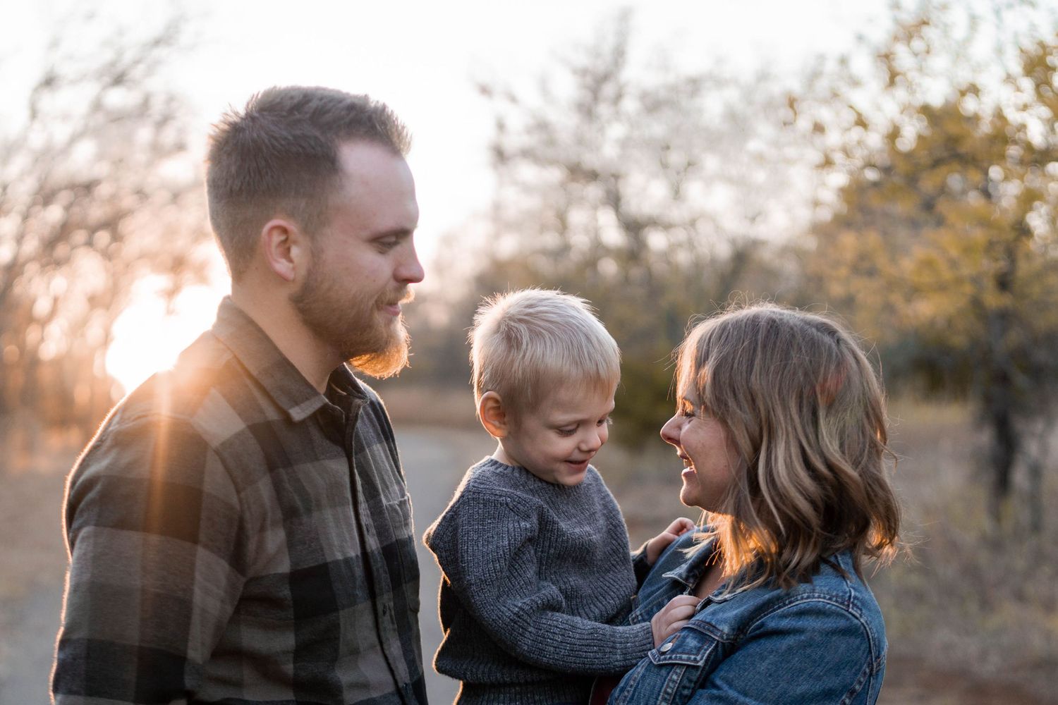 A young family shares a tender moment outdoors at golden hour, with parents gazing lovingly at their toddler son.
