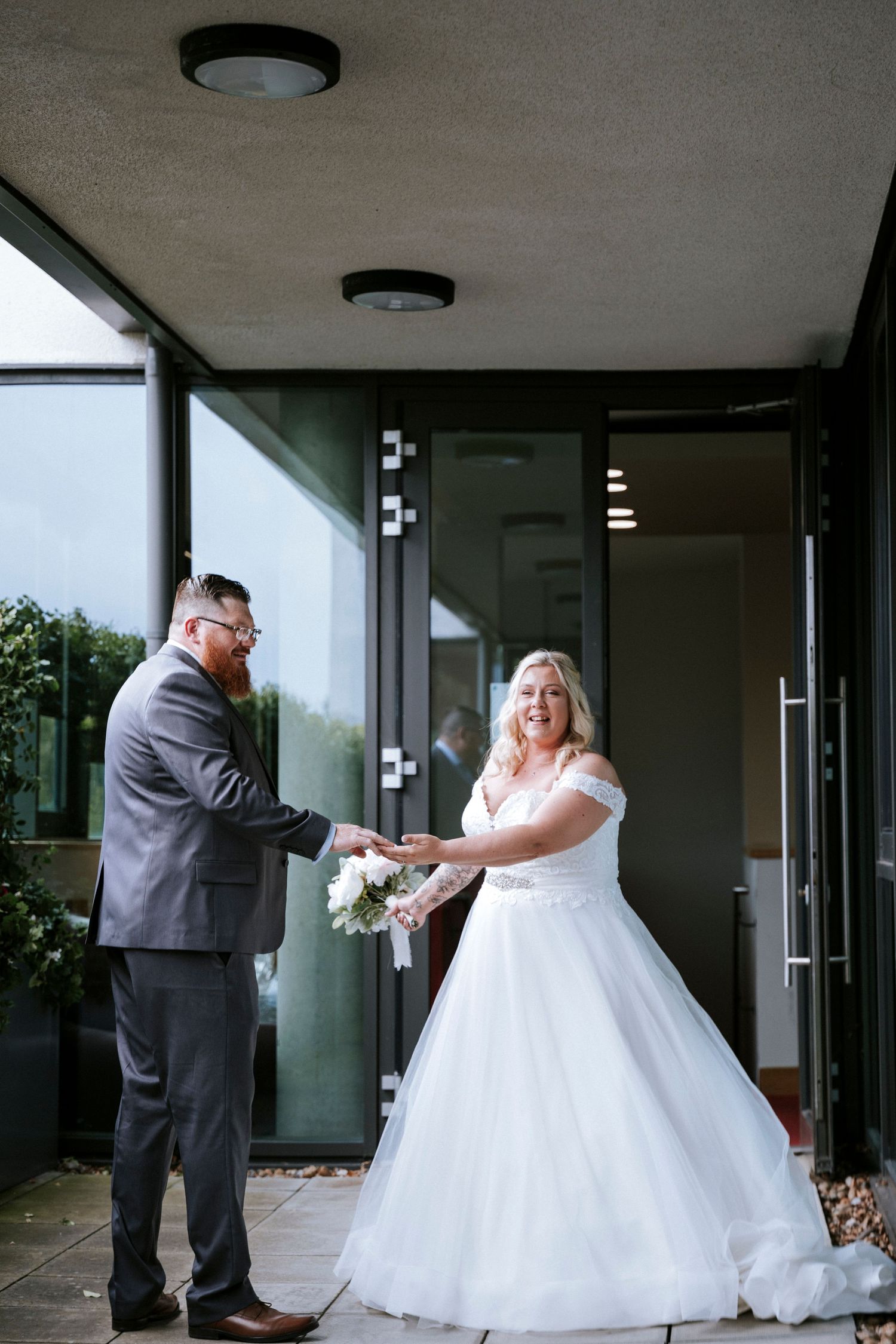 A newlywed couple shares a moment at the entrance of a modern building with glass doors.