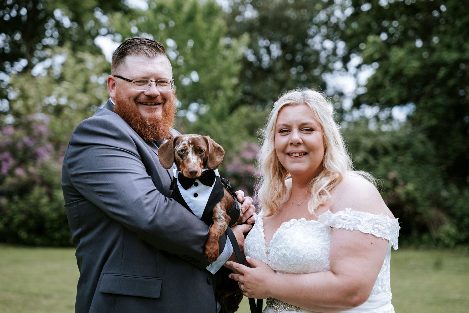 A happy wedding couple holds their small brown dog during outdoor portraits.