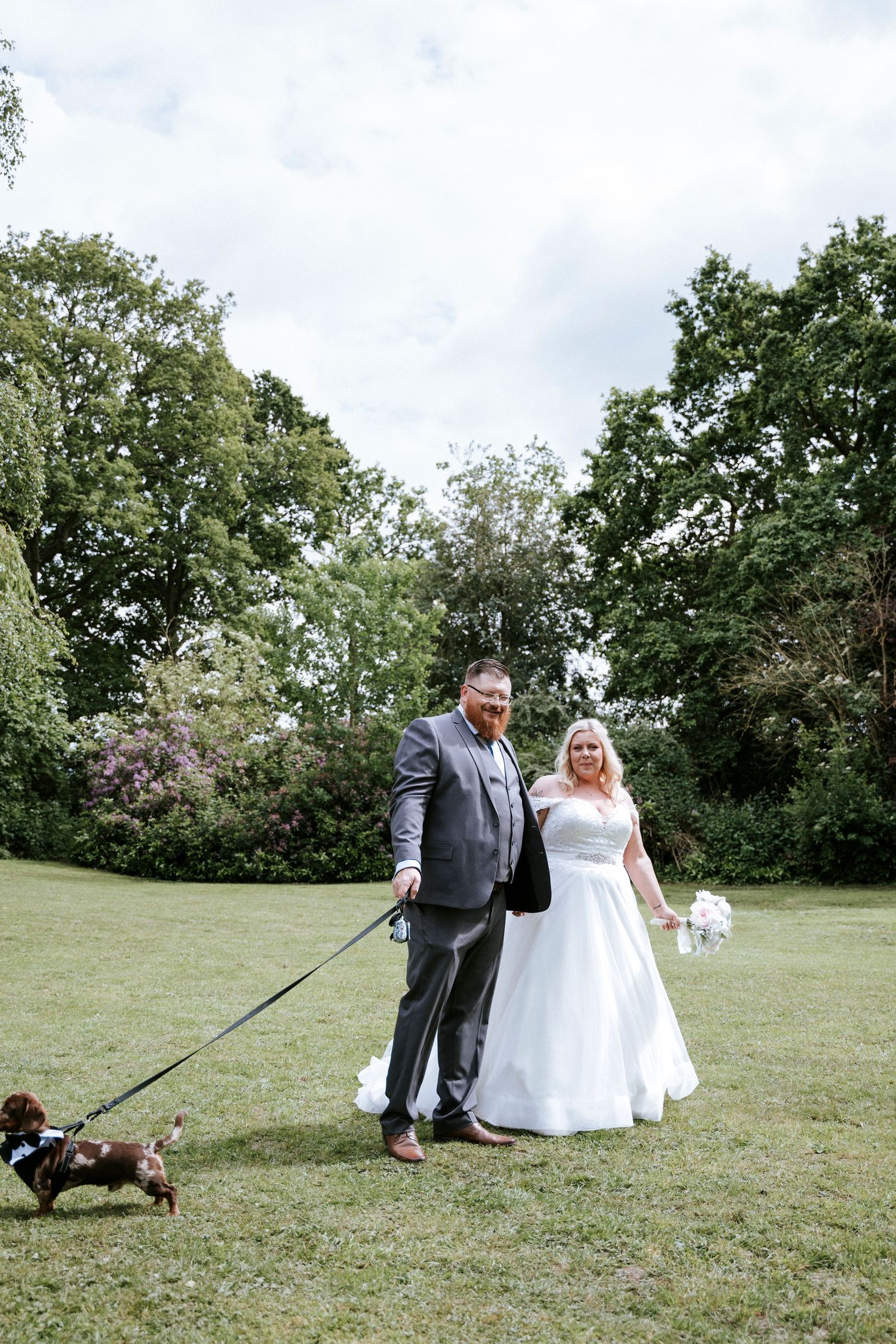 A couple poses on a green lawn while walking their small dog on a leash.