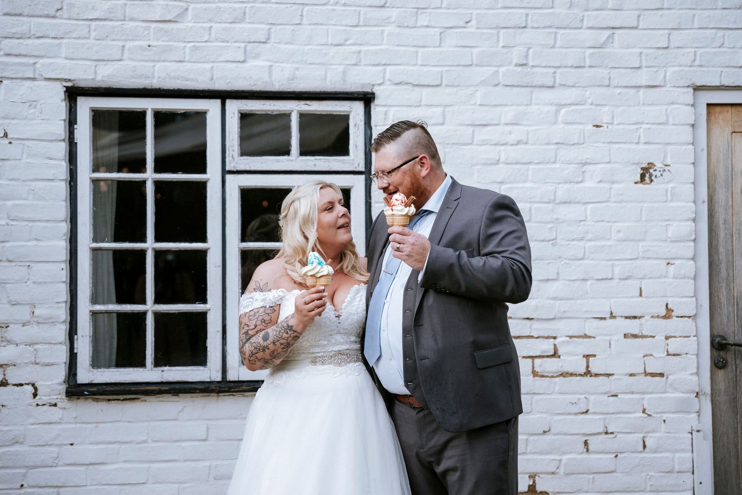 A couple shares a laugh against a white brick wall with a window.