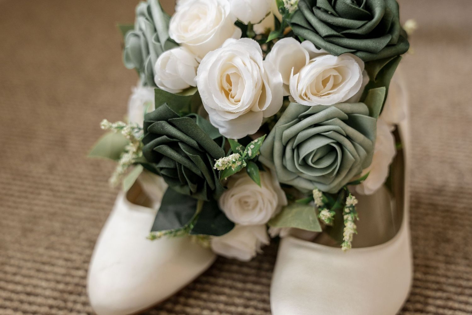 White and sage green roses arranged in a delicate bridal bouquet next to white wedding shoes.