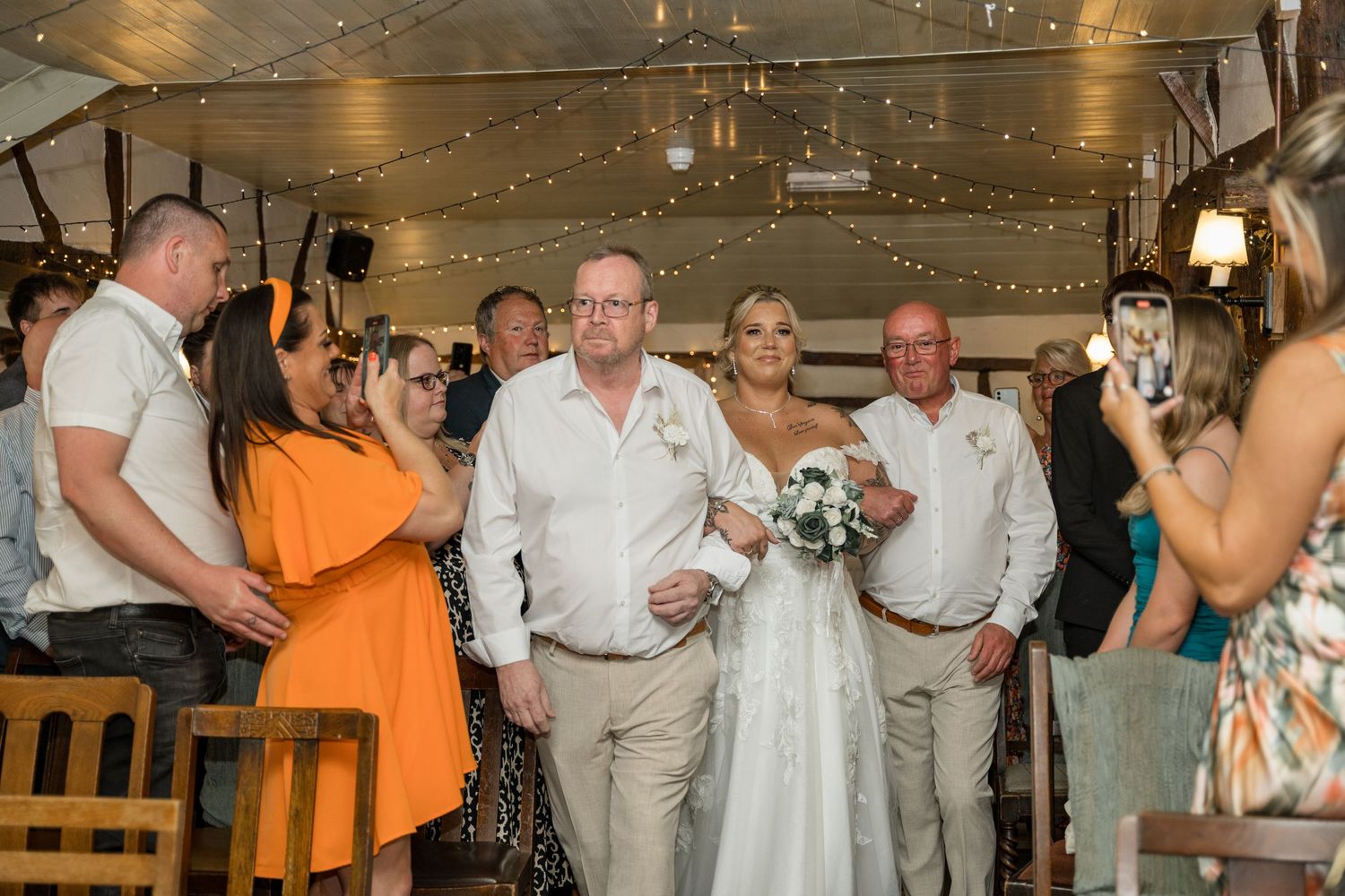 Wedding party and guests gather inside a rustic venue decorated with hanging lights for celebration.