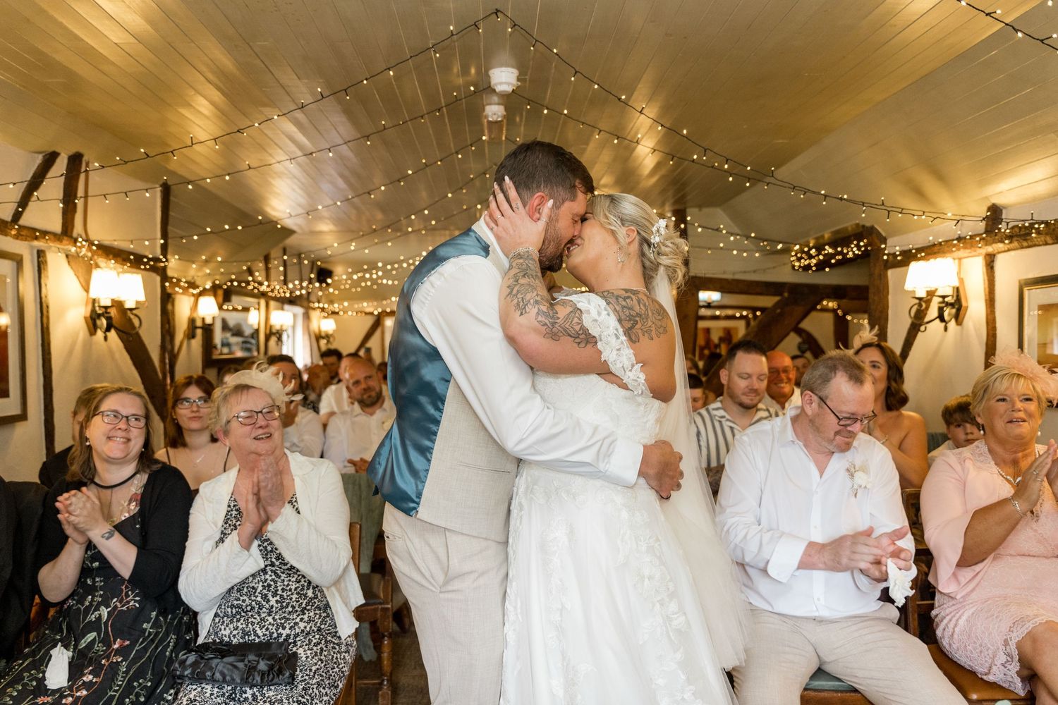 Wedding guests celebrate and applaud during a reception in a venue decorated with string lights.