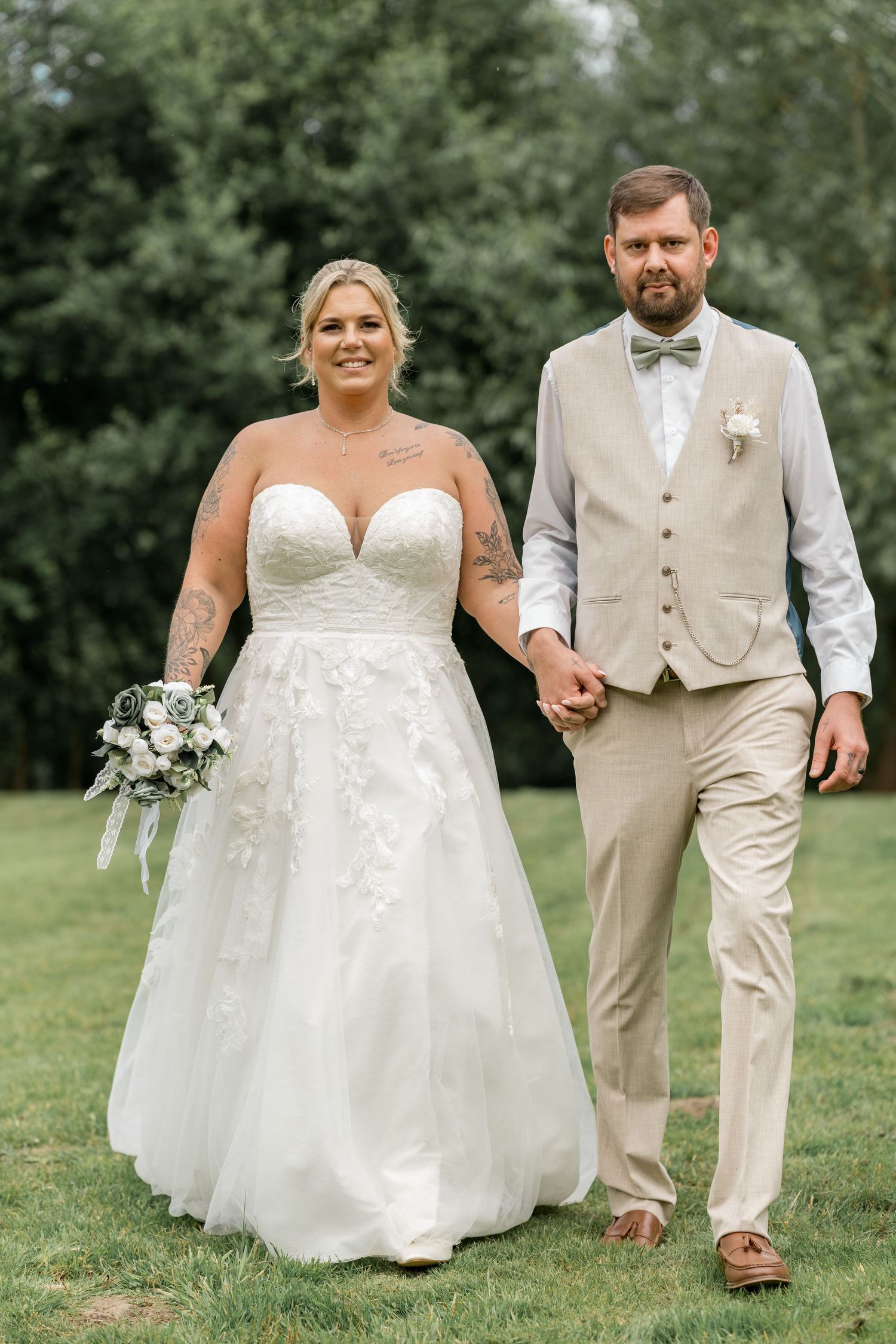A couple poses together outdoors at their wedding, with the bride in a lace gown and groom in a tan suit.