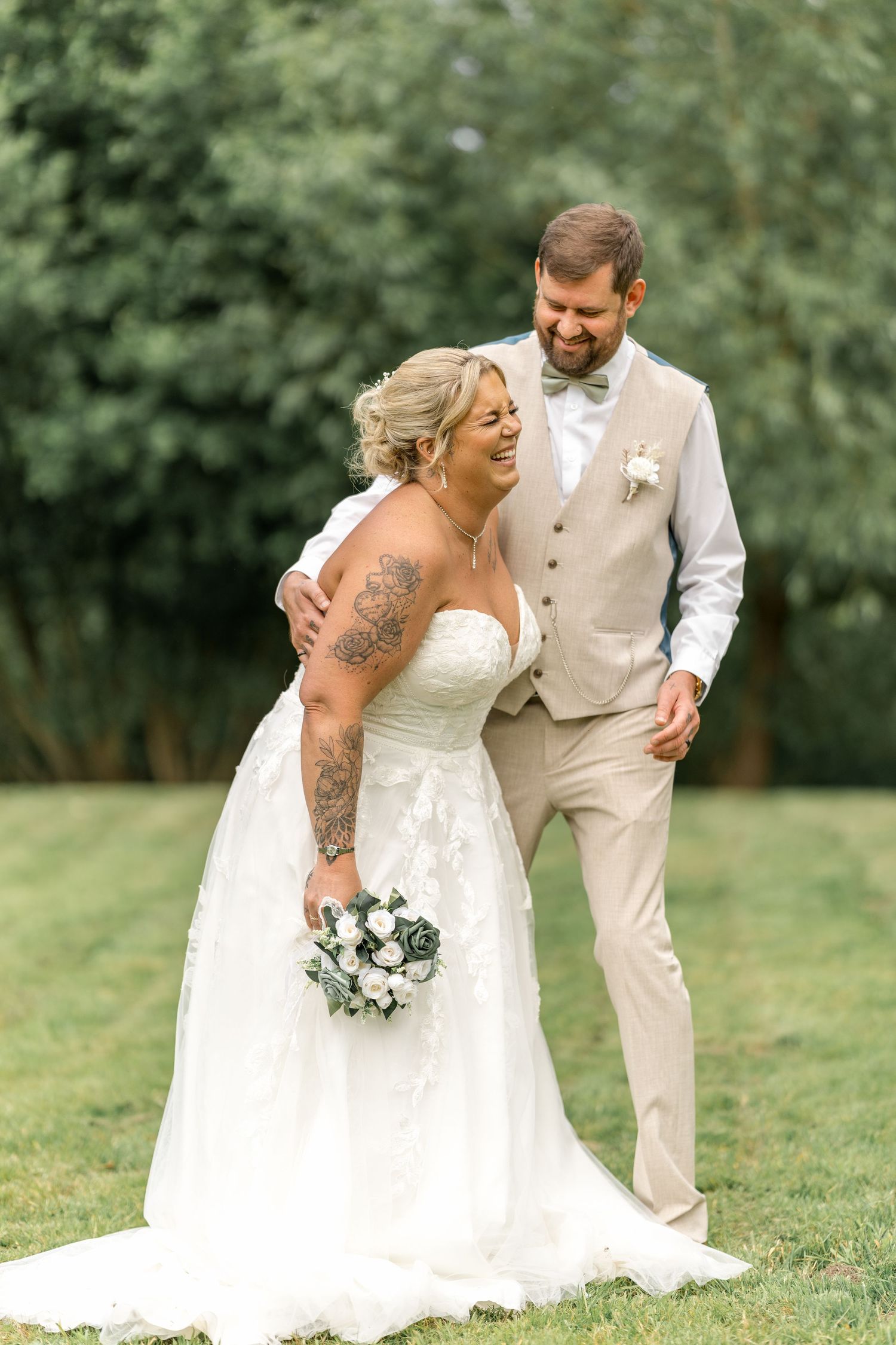 A happy wedding couple shares a tender moment on grass with a bouquet of white flowers.