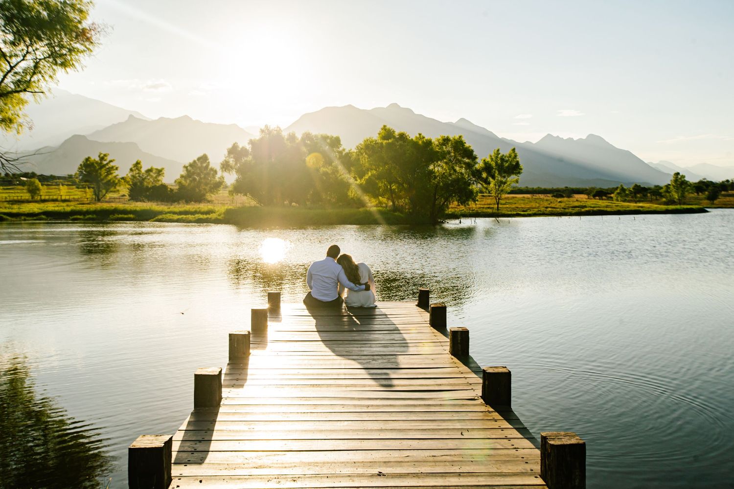 A couple sits on a wooden pier overlooking a serene lake and mountains at sunset.