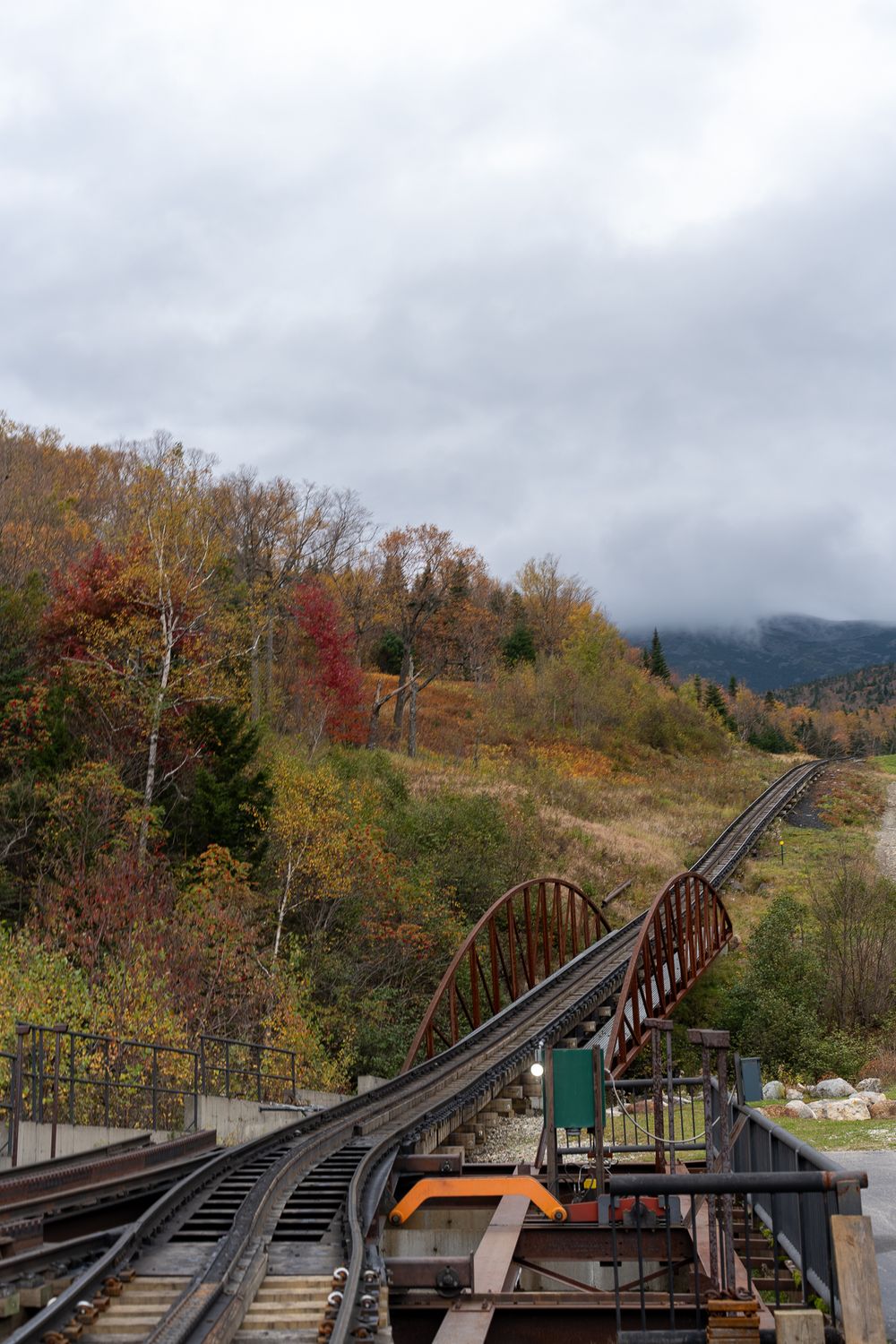 A scenic railway track crosses a rustic bridge amid vibrant fall foliage on a cloudy autumn day.